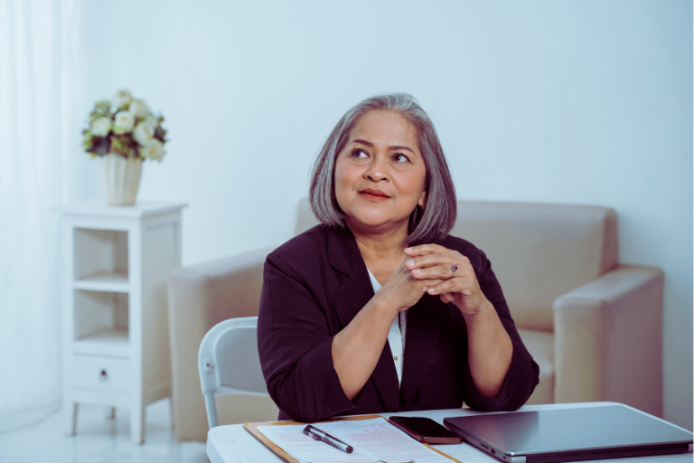 older woman thinking at desk with laptop and cell phone