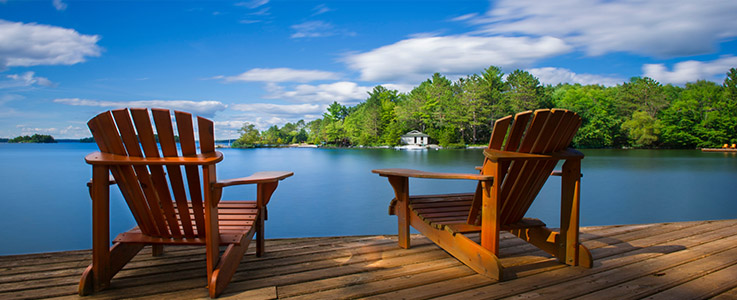 Two Muskoka chairs sitting on a wood dock facing a calm lake. Ac